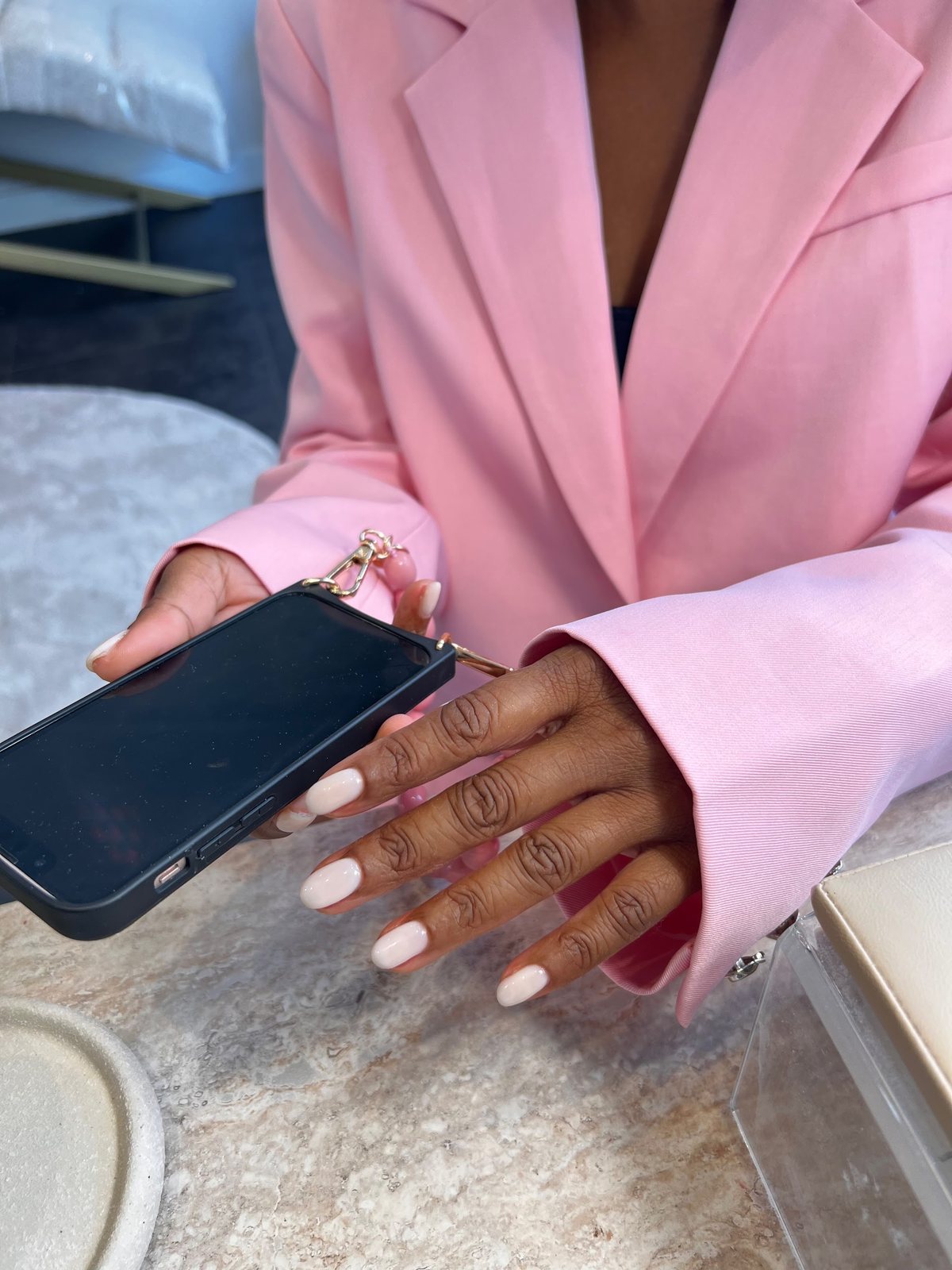 Handen met melkwitte nagels die een telefoon vasthouden, roze blazer